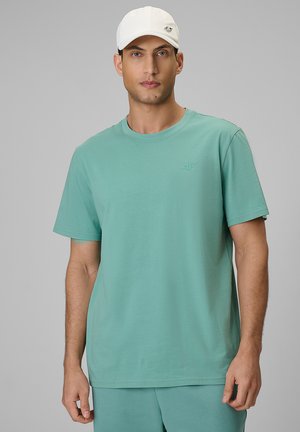 Mężczyzna ubrany w jasnoturkusowy t-shirt z krótkim rękawem i pasujące szorty oraz białą czapkę z daszkiem na szarym tle.