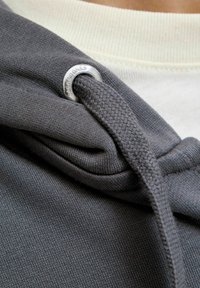 Material de sudadera gris con una capucha texturizada. Presenta un ojal plateado para el cordón, que es tejido y detallado. Fondo claro.