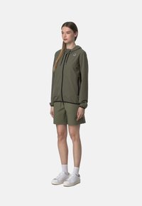 Giacca a zip verde oliva con cappuccio, dettagli neri e tasche, abbinata a pantaloni corti coordinati. Il modello indossa sneakers bianche e calzini.