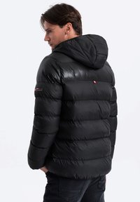 Schwarze gepolsterte Jacke mit Kapuze, mit einer glänzenden Außenseite, horizontalen Nähten und einem kleinen Logo am Ärmel. Verstellbare Ärmelbündchen.