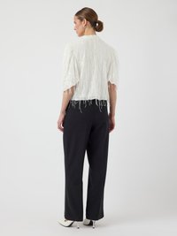 Witte textuurjacket met franjes en korte mouwen, gecombineerd met zwarte wijde broek en puntige witte schoenen.
