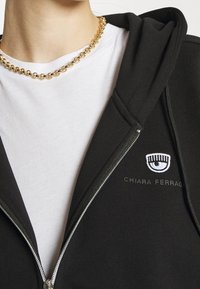 Musta vetoketjullinen huppari, jossa vasemmalla puolella valkoinen silmää ja tekstiä esittävä logo, päällä valkoinen T-paita, teksturoitu kangas. Kultainen ketjukaulakoru.