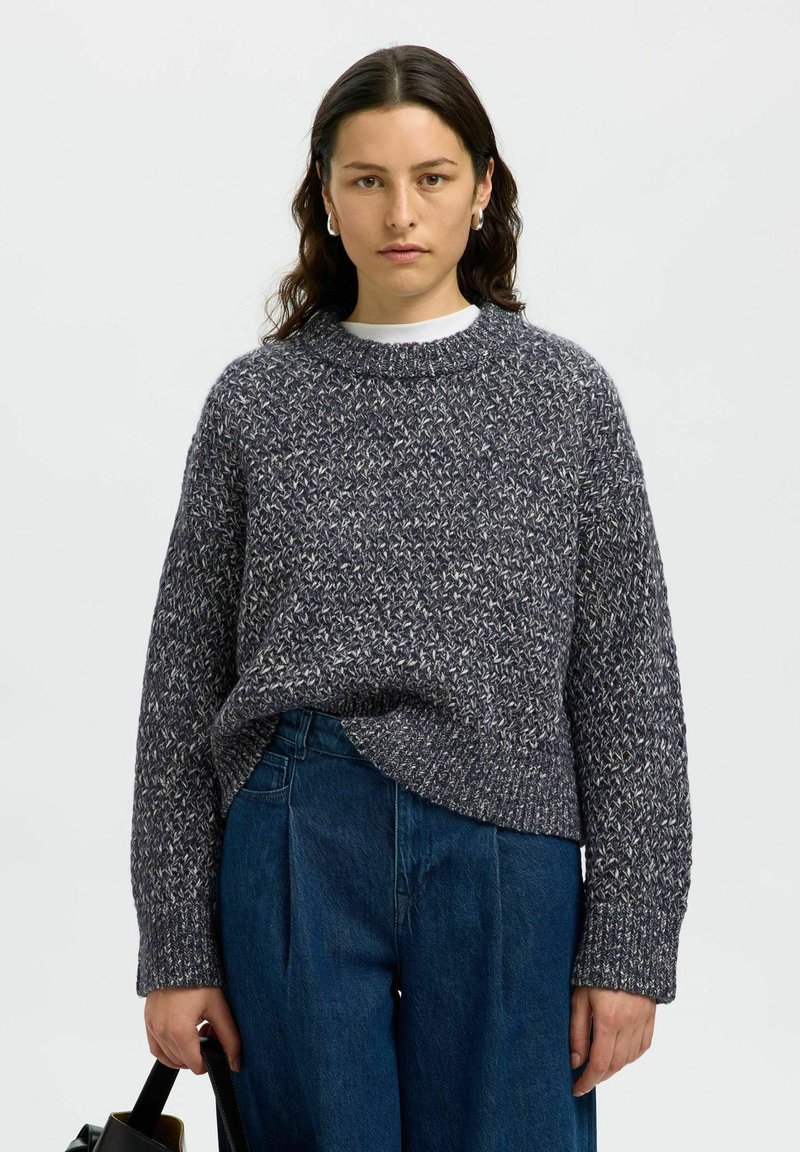 Sweter dzianinowy w ciemnoszarym kolorze z teksturowanym wzorem. Posiada okrągły dekolt i luźny krój, zestawiony z niebieskimi spodniami typu wide-leg. Prosty styl.