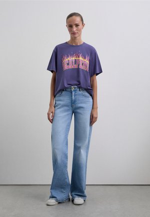 Paarse oversized T-shirt met vlamgrafiek en de tekst "SCLPER", gecombineerd met lichtblauwe wijde jeans en witte sneakers. Soepele, casual stof.