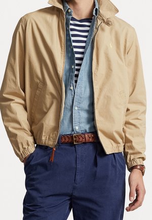 Homme portant une veste beige sur une chemise en denim bleu et un t-shirt rayé bleu marine, accompagné d'une ceinture tressée marron et d'un pantalon bleu marine.