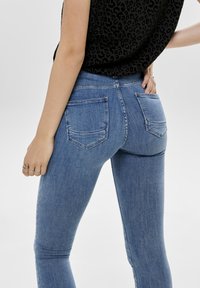 Vaqueros de denim azul ajustados con un ligero estiramiento, que presentan bolsillos traseros y una textura suave, ligeramente desteñida. Combinados con una blusa negra.