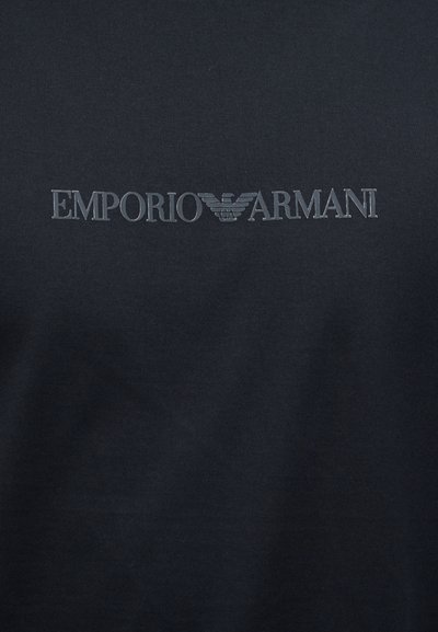 Μαύρο ύφασμα με το κεντρικό λογότυπο Emporio Armani σε σκούρο γκρι, που περιλαμβάνει το όνομα της μάρκας και το εμβληματικό σύμβολο αετού.