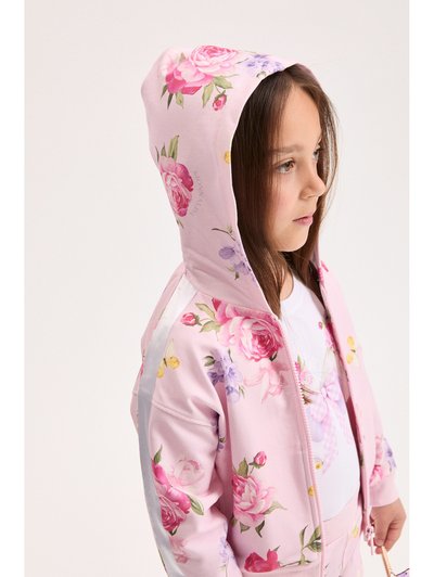 Νεαρή κοπέλα που κοιτάζει δεξιά, φορώντας ροζ φλοράλ φούτερ με φερμουάρ και κουκούλα καλυμμένη στο κεφάλι, καθώς και ταιριαστό παντελόνι, σε απλό φωτεινό φόντο.