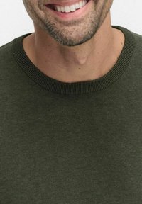 Tumma vihreä neulepusero, jossa on ribbireunainen pyöreä kaula-aukko ja sileä pinta, paljastaen osan kaulasta ja leuasta sen alla.