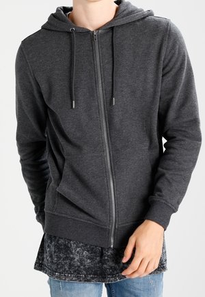Person trägt einen dunkelgrauen Kapuzenpullover mit Reißverschluss und Kordeln über einem schwarzen Acid-Wash-Shirt und blauen Jeans, steht vor einem einfarbigen Hintergrund.