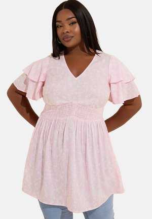 Kobieta w jasnoróżowej bluzce w kwiaty z dekoltem w kształcie litery V, z marszczonym pasem i falbanianymi, krótkimi rękawami, stojąca z rękami na biodrach na białym tle.
