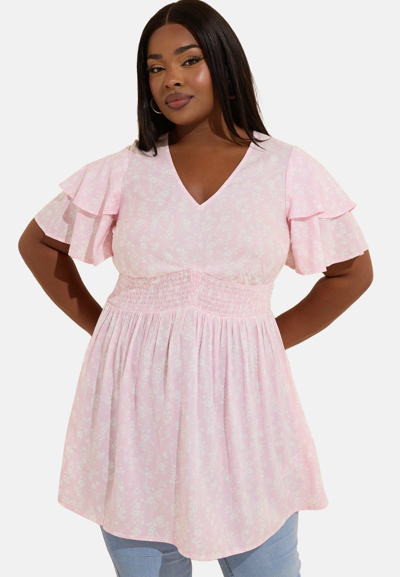 Mujer con una blusa de flores rosa claro con escote en V, cintura fruncida y mangas cortas con volantes, de pie con las manos en las caderas contra un fondo blanco.