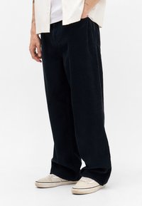 Mand iført løse sorte fløjlsbukser, beige sneakers, hvid T-shirt og cremet kortærmet skjorte med hånden i lommen.