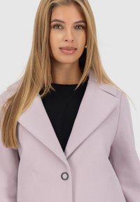 Giovane donna con capelli biondi lisci che indossa un cappotto lilla chiaro sopra una maglietta nera, di fronte con un'espressione neutra.