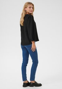 Black, textured long-sleeve top with puffed shoulders, paired with fitted blue jeans and black shoes. Model viewed from the side.