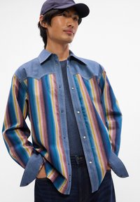 Τζιν πουκάμισο με πολύχρωμες κάθετες ρίγες, ανοιχτό μπλε γιακά, κουμπιά με κούμπωμα, και μακριά μανίκια. Φορεμένο πάνω από μπλε ναυτικό μπλουζάκι και σκούρους τζιν.