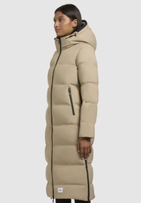 Beige Daunenjacke mit Kapuze, langer Schnitt, vertikaler Steppung und mit Reißverschlüssen versehenen Taschen. Glatte Textur und leichtes Material.