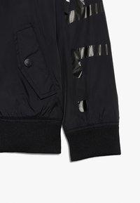 Musta bomber-takki, jossa on kiiltävä pinta, vetoketjullinen tasku vasemmassa hihassa ja ribbauskaihtimet, joissa on leikattuja korostuskuvioita.