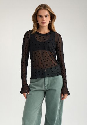 Jeune femme portant un haut noir transparent à manches longues à pois sur un débardeur noir et un pantalon vert clair taille haute à jambes larges, debout devant un fond uni.