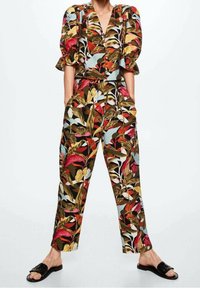 Donna che indossa una blusa a maniche a sbuffo con stampa floreale vivace e pantaloni a vita alta abbinati, con le mani nelle tasche, abbinati a sandali neri.