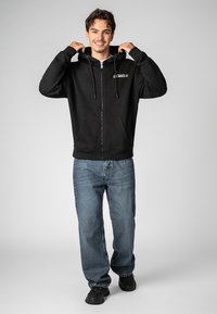 Black zip-up hoodie with drawstrings and front pockets, worn over blue jeans; features a logo on the left chest. Textured fabric.