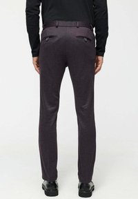 Man wearing dark purple tailored pants with back buttoned pockets, black shoes, and a black long-sleeve top, standing against white background.