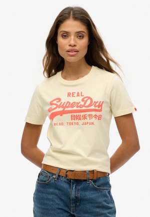 Cream-colored cotton t-shirt with red graphic text reading "REAL SuperDry," alongside Japanese characters, worn with blue jeans and brown belt.