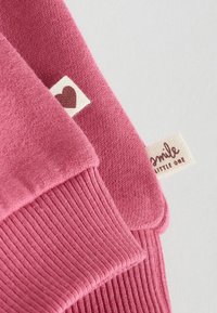 Rosa stickad tyg med ribbat mönster och två etiketter: en med ett hjärtformigt motiv och en annan med texten "leks litet barn."
