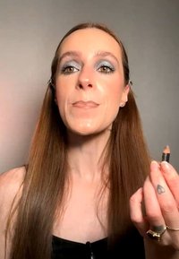Femme aux longs cheveux bruns et maquillage argenté aux yeux tenant un crayon à sourcils marron près de son visage sur un fond uni.