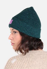 Bonnet en tricot vert avec une texture côtelée et une étiquette rose. Le modèle porte un pull beige, mettant en valeur le design ajusté du bonnet.