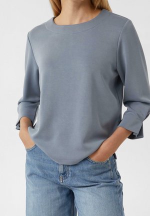 Kobieta ubrana w niebiesko-szarą bluzkę z długim rękawem i okrągłym dekoltem oraz jasnoniebieskie dżinsy, z rękami w kieszeniach, na jednolitym tle.