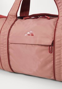 Sac de sport en nylon rose avec des accents rouges, doté d'une poche zippée, de doubles poignées et d'un emblème de logo brillant. Texture lisse et design structuré.