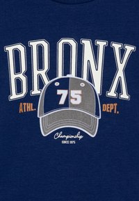 Maglietta blu navy con grande testo bianco "BRONX", grafica di un cappellino grigio e blu con il numero "75", e testo bianco "Championship since 1975" sottostante.