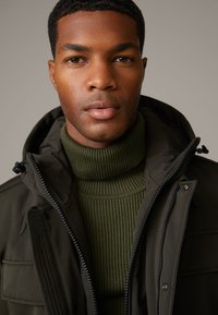 Dunkelgrüne Kapuzenjacke mit Reißverschluss und Taschen, getragen über einem gerippten, olivgrünen Rollkragenpullover. Glatte Texturen und praktisches Design.