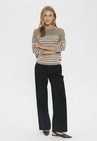Donna con le braccia incrociate, che indossa un maglione a righe taupe e crema, pantaloni neri a gamba larga e décolleté neri su uno sfondo bianco.