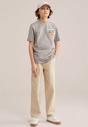 Fant, ki nosi sivo majico z grafiko pomarančnega sadja, bež hlače, bele superge in bež kapo, stoji z eno roko v žepu.