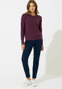 Maglione viola a maglia con un motivo testurizzato, accompagnato da pantaloni blu scuro e sneaker bianche, esibito in una posa eretta.