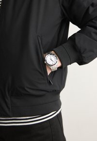 Veste bomber noire avec une poche zippée, dotée de poignets côtelés et d'un ourlet rayé. Une montre argentée avec un cadran blanc est visible au poignet.
