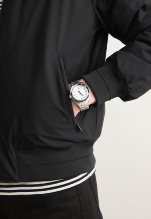 Schwarze Bomberjacke mit einer Reißverschlusstasche, gerippten Bündchen und einem gestreiften Saum. Eine silberne Uhr mit einem weißen Ziffernblatt ist am Handgelenk sichtbar.