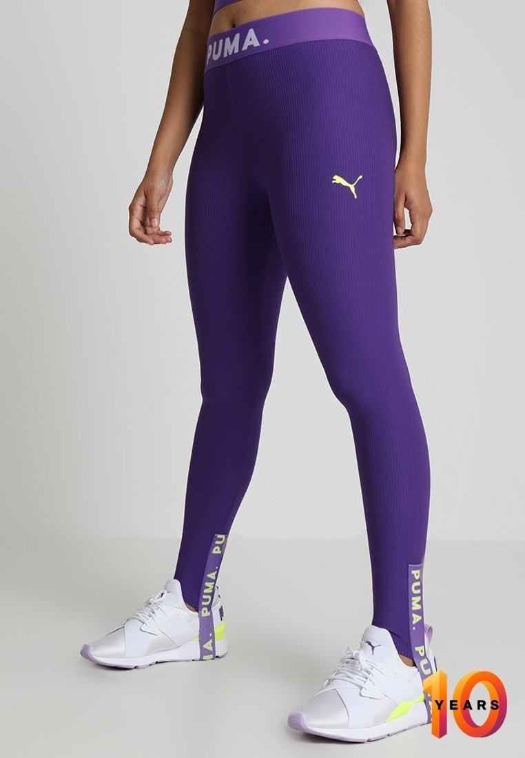 Fioletowe legginsy z prążkowanym wzorem z żółtym logo Pumy, pasem z napisem "PUMA" oraz kontrastowym tekstem na kostce. W komplecie z białymi sneakersami.