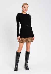 Donna che indossa un miniabito nero a maniche lunghe con un orlo in stampa leopardata e stivali di pelle neri fino al ginocchio, in piedi su uno sfondo bianco.