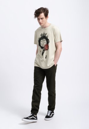 T-shirt in cotone beige con stampa grafica raffigurante un design con punto interrogativo. Abbinato a pantaloni cargo verdi scuri e sneakers nere.