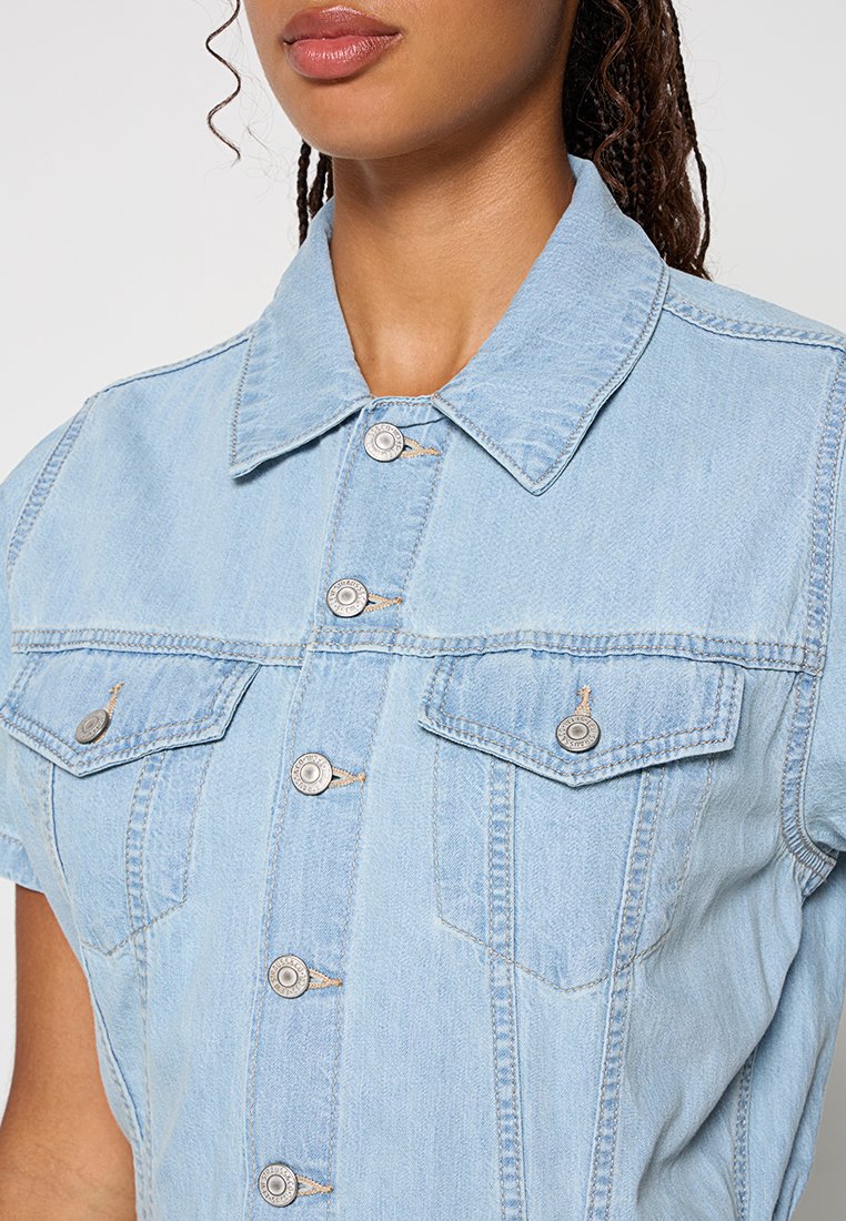 Chemise en denim bleu clair à manches courtes, dotée d'un col pointu, de boutons-pression à l'avant et de deux poches poitrine avec des accents métalliques.