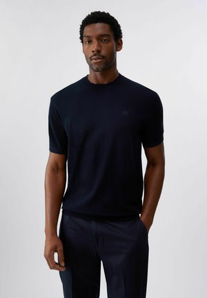 Uomo con capelli corti e ricci che indossa una maglietta nera a maniche corte e pantaloni scuri, in piedi contro uno sfondo chiaro e uniforme.