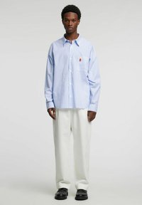 Stripete skjorte med knapper i lyseblått og hvitt, med en lomme som har en liten brodert detalj. Kombinert med løse hvite bukser og svarte sko.