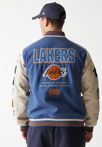 Blue and beige varsity jacket featuring "LAKERS" embroidered on the back, with logos and patterns on sleeves. Ribbed collar and waistband.