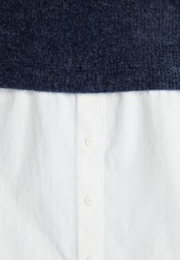 Haut en maille bleu marine avec un motif texturé au-dessus d'une chemise blanche lisse, présentant un placket à boutons et six boutons blancs sur le devant.
