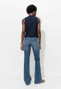 Haut sans manches en marine associé à un jean évasé bleu avec des poches arrière. Modèle portant des chaussures à imprimé animal et un bracelet en or, vu de dos.