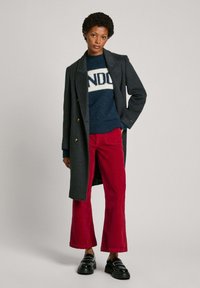 Manteau en laine gris avec un design croisé, porté sur un pull bleu marine avec un imprimé "NDO", associé à un pantalon évasé en velours côtelé rouge et des chaussures noires.