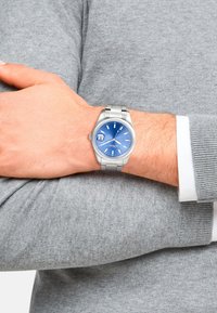Edelstahlarmbanduhr mit blauem Zifferblatt, silbernen Stundenmarkierungen und einem Datumsanzeige. Am Handgelenk getragen, kombiniert mit einem grauen Pullover.